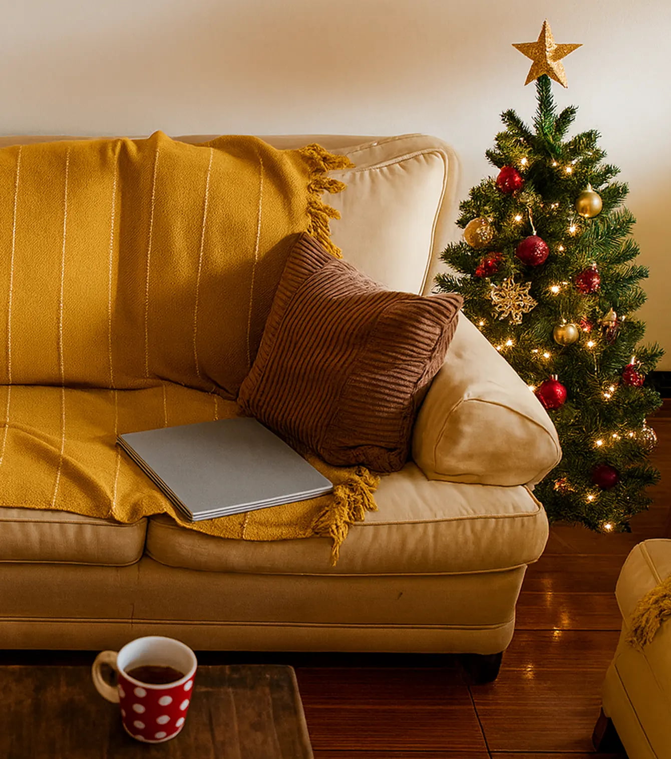 Mysig beige soffa med gul filt och kudde, en laptop och kaffekopp på bordet, och en julgran i bakgrunden.